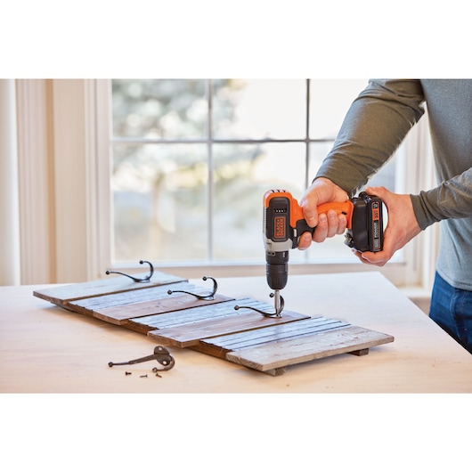 BLACKDECKER BCD702C1 cordless drill in use attaching metal hooks to a wooden board on a table indoors.