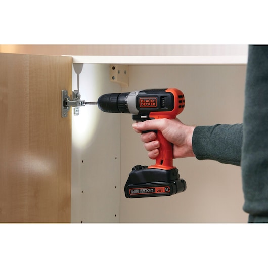 BLACK+DECKER BCD702C1 cordless drill being used to install cabinet hinge, orange and black with LED light.