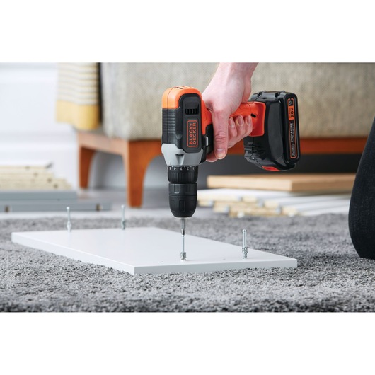 Photo of BLACK+DECKER BCD702C1 cordless drill driving a screw into a white board during furniture assembly.