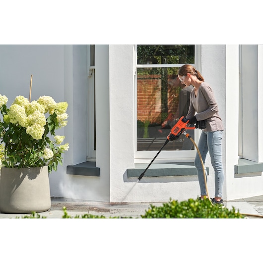 Photo of person using BLACKDECKER BCPC20D1 orange power washer to clean patio near white house and potted flowers.