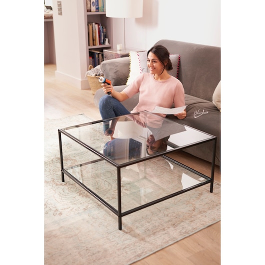 Photo of person using BLACKDECKER BCRTA601I tool while sitting on grey sofa in living room with glass coffee table.