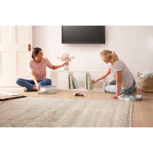 BLACKDECKER BCRTA601I cordless screwdriver on floor between two women assembling a white shelf in a living room.