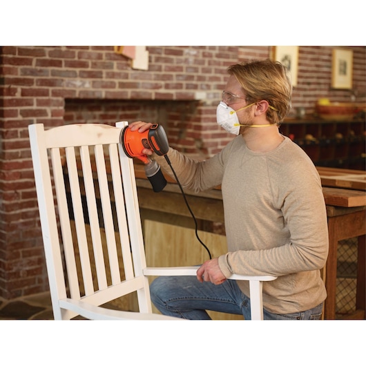 Person sanding a white wooden chair with BLACKDECKER BDERO100 electric sander in a workshop.