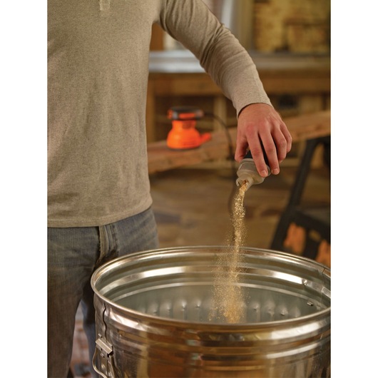 Photo of person emptying dust from BLACKDECKER BDERO100 sander container into a metal trash can in workshop.