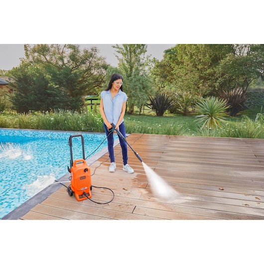 Photo of BLACKDECKER BEPW1800T orange pressure washer cleaning wooden deck by a pool in a garden setting.