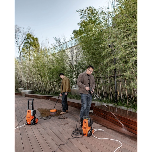 Two people using BLACKDECKER BEPWB1740 pressure washers to clean wooden deck outdoors, both units visible on the ground.