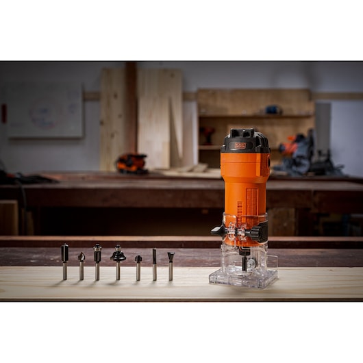 A BLACK+DECKER router tool in orange color is placed on a wooden workbench, with seven different router bits lined up in front of it. The background shows a workshop setting with tools and wood panels.