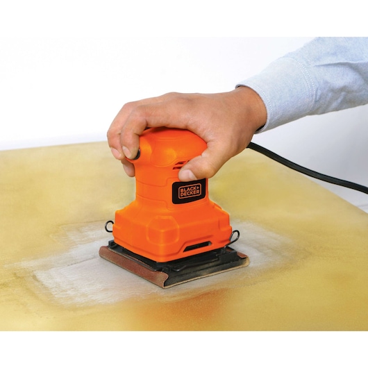 BLACK+DECKER BS200 orange sheet sander in use on wood surface, close-up hand holding tool.