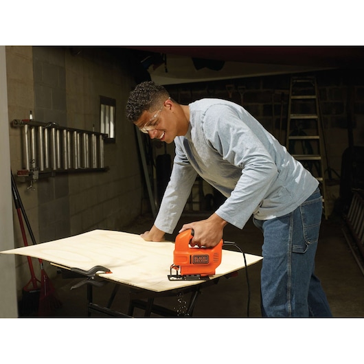 Photo of a person using a BLACK+DECKER KS501 jigsaw to cut plywood on a workbench in a garage.