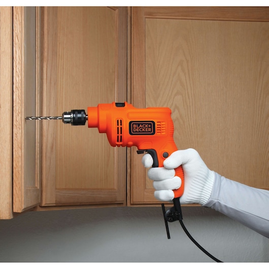 Photo of BLACK+DECKER TP555 orange corded drill held by gloved hand, drilling into wooden cabinet.